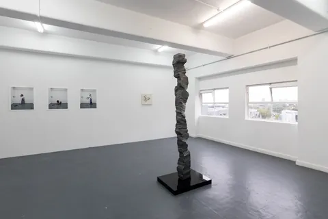A tall dark aluminium column on a black plinth in a white gallery with windows