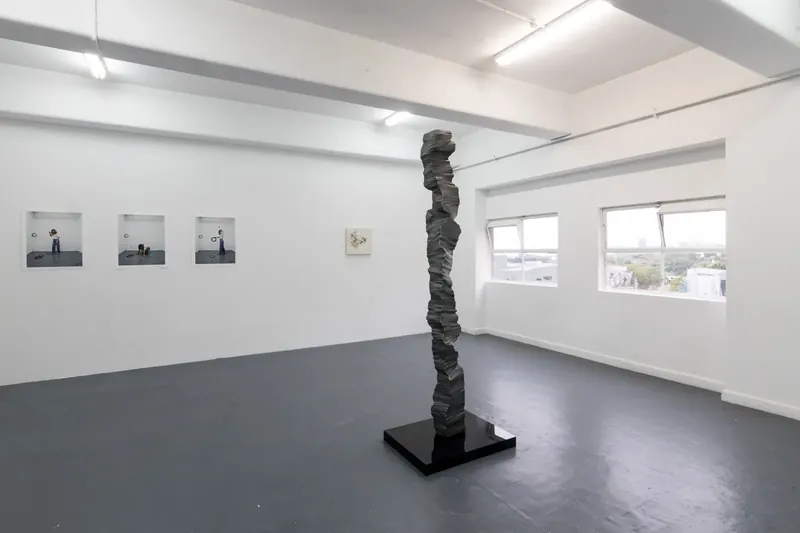 A tall dark aluminium column on a black plinth in a white gallery with windows