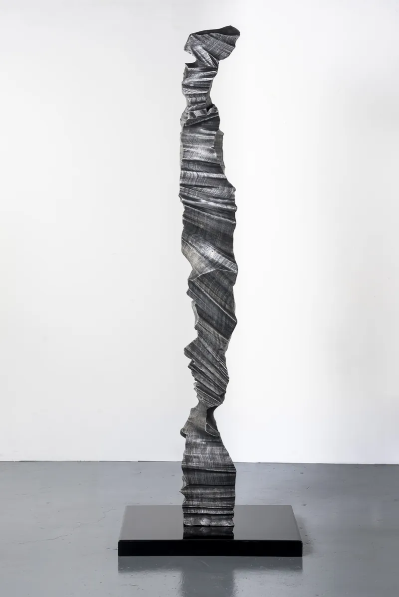 A tall cast aluminium column with a folded, layered surface, on a black plinth.