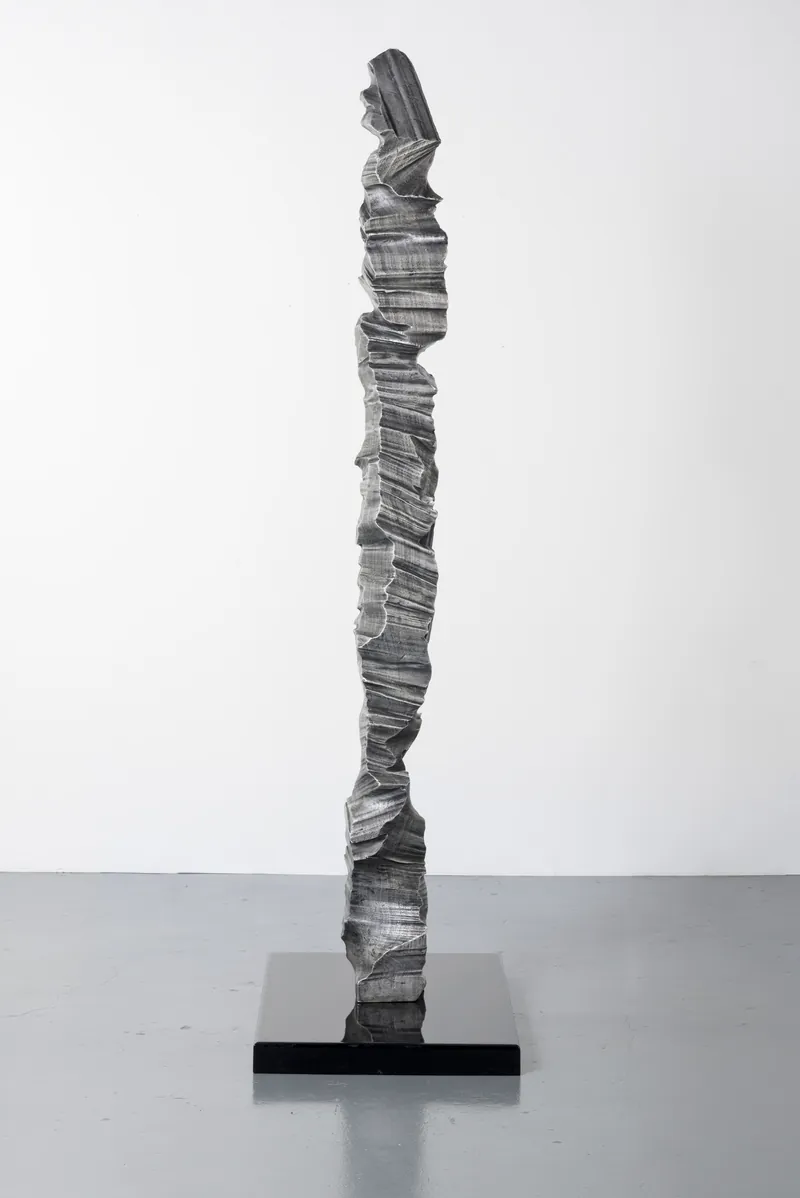A tall, narrow aluminium column on a black plinth, full view
