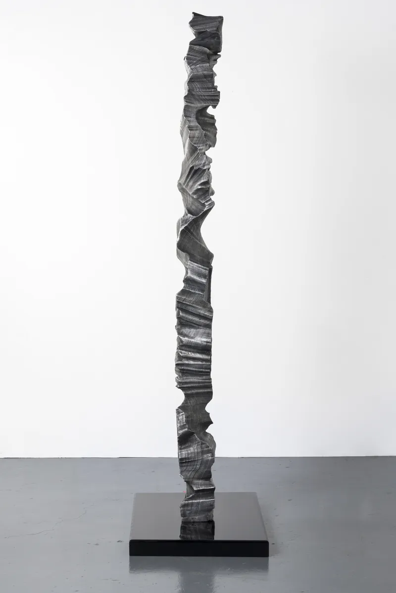 A single slender aluminium column on a black plinth against a white wall, full view